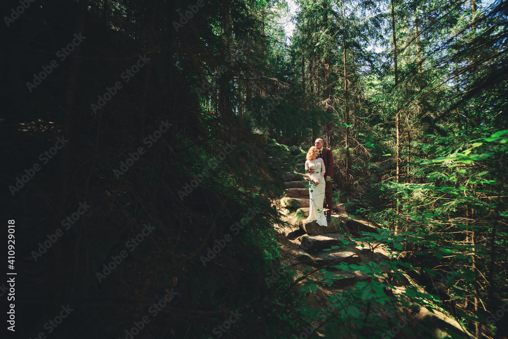 Happy stylish couple newlyweds in the green forest on summer day. bride in long white dress and groom in red suit are hugging. wedding day.
