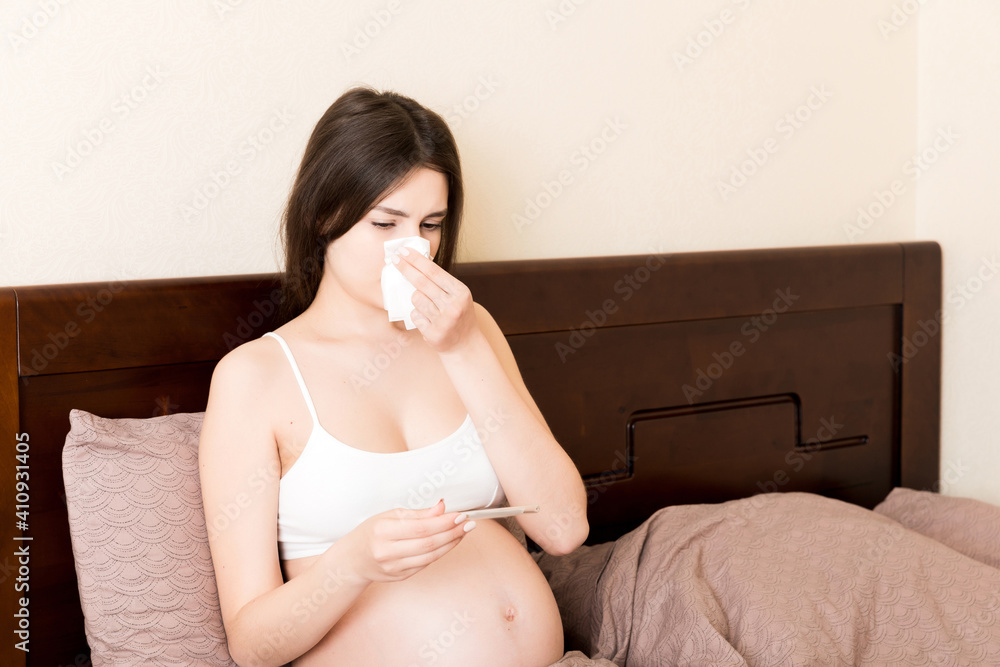 Portrait of young unhappy pregnant woman leaning on pillows on bed and blowing her nose into tissue. Future mom caught cold and sneezing