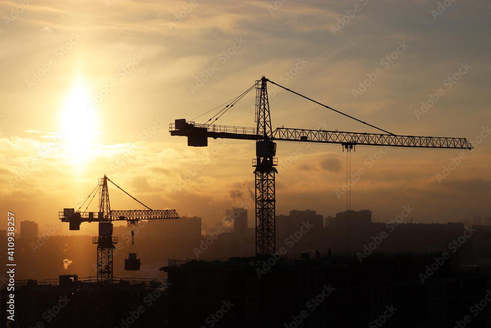 Silhouettes of construction cranes and unfinished residential building during snow on sunrise background. Housing construction, apartment block in winter city