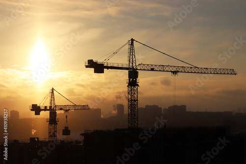 Silhouettes of construction cranes and unfinished residential building during snow on sunrise background. Housing construction, apartment block in winter city