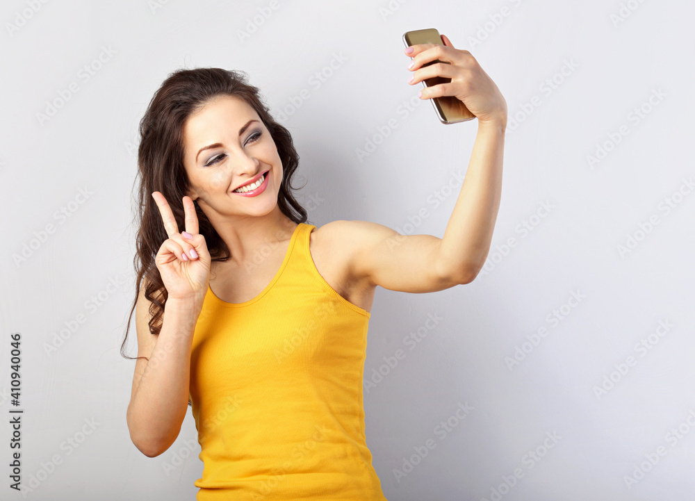 Funny positive smiling woman with long hair making self photo on mobile phone and showing victory sing by hand in yellow bright t-shirt on light blue background Funny positive smiling woman with long hair making self photo on mobile phone and showing victory sing by hand in yellow bright t-shirt on light blue background