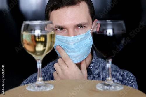 Choosing alcohol, man in medical face mask looks at the glasses with white and red wine. Concept of tasting and party during coronavirus pandemic