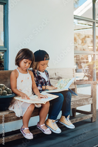 Two concentrated kids dressed in casual clothing sit on a park bench and read an interesting book in datyime. photo