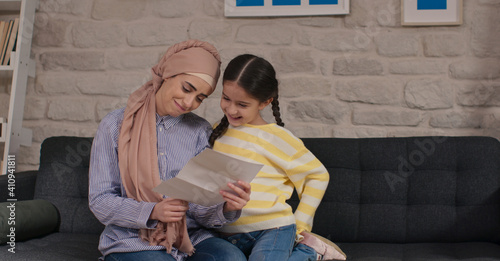 Headscarved mother and daughter are sitting on the sofa and reading letters together. The mother is upset by the news in the letter. 