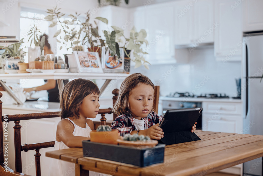 Happy family. Portrait of a brother and sister which use a tablet sitting at table in the kitchen of modern apartment.