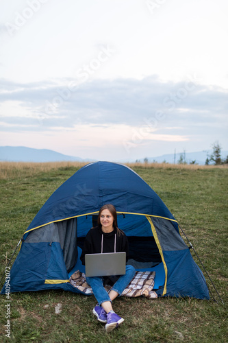 Woman working on laptop in tent in nature. Young freelancer sitting in camp. Relaxing in camping site in mountains. Remote work, outdoor activity in summer. Happy girl relaxing, work on vacation