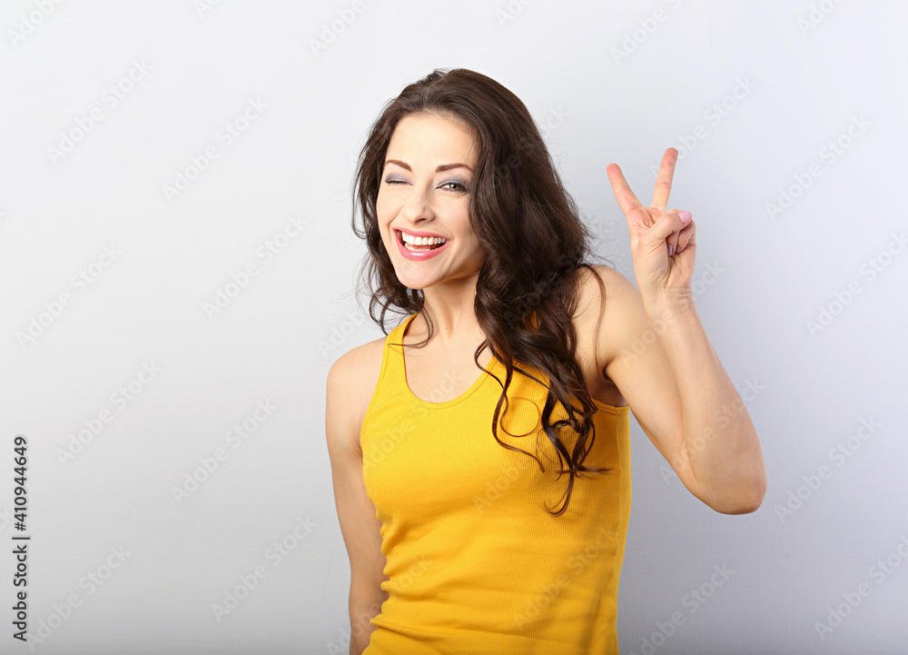 Beautiful smiling woman showing two fingers victory sign and winking one eye in casual yellow t-shirt clothing on blue background with empty copy space. Beautiful smiling woman showing two fingers victory sign and winking one eye in casual yellow t-shirt clothing on blue background with empty copy space.