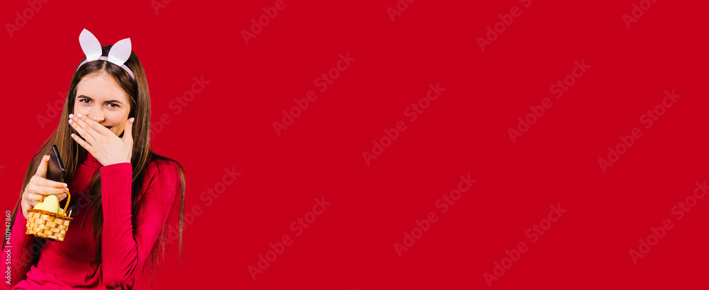 Banner,long format. Happy girl with ears covers her smile with her hand and holds decorative Easter eggs on a red background with empty advertising space. Banner,long format. Happy girl with ears covers her smile with her hand and holds decorative Easter eggs on a red background with empty advertising space.