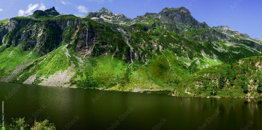 rocky mountains with waterfalls and green plants at a lake panorama
