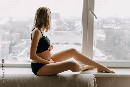 Pregnant woman in black sport underwear, sitting on windowsill by the window at home.