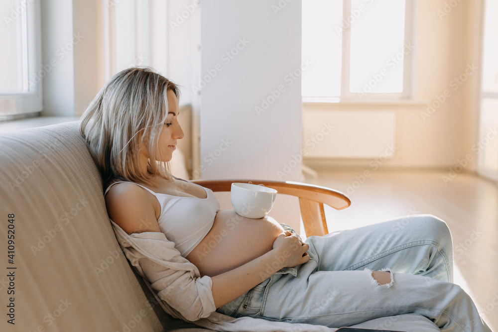 Pregnant woman sitting on a sofa at home with a white cup of coffee or tea on her belly. Pregnant woman sitting on a sofa at home with a white cup of coffee or tea on her belly.