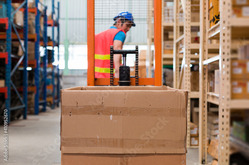 Asian male warehouse worker in safety vest and helmet driving and operating on forklift truck in the industry storage warehouse with shelf pallet automotive spare parts parcel blur background
