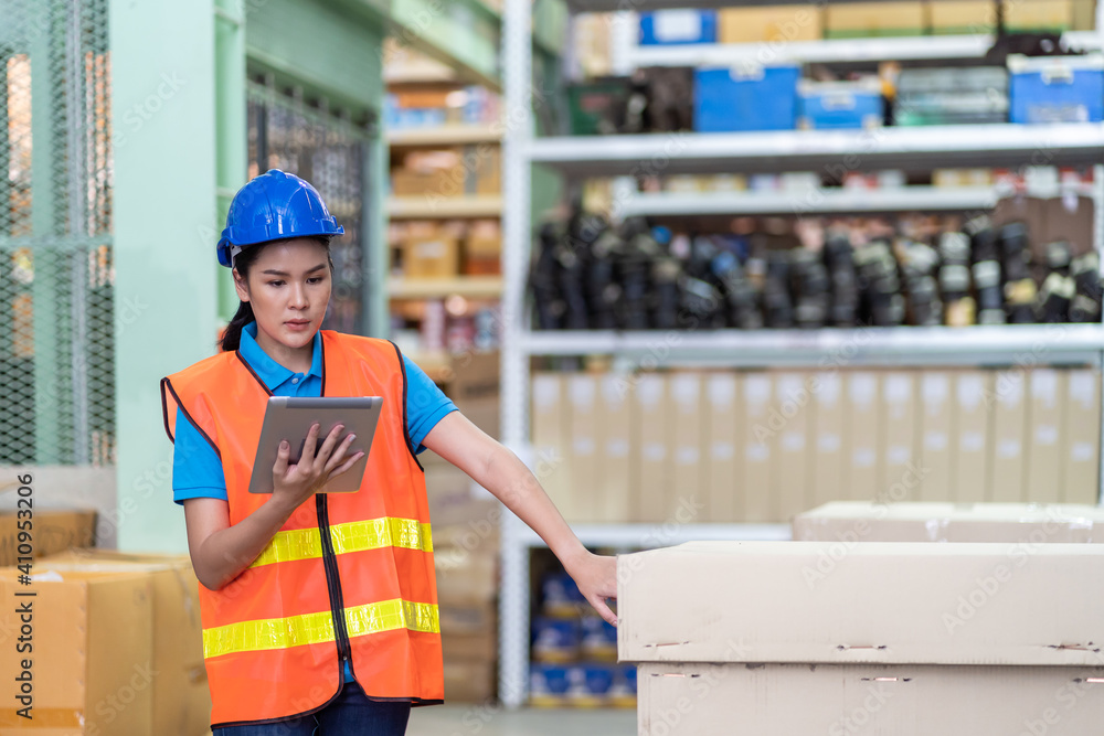Asian female worker working with digital tablet checking products in parcel box at spare parts warehouse. Woman wearing safety vest and helmet working at warehouse Asian female worker working with digital tablet checking products in parcel box at spare parts warehouse. Woman wearing safety vest and helmet working at warehouse