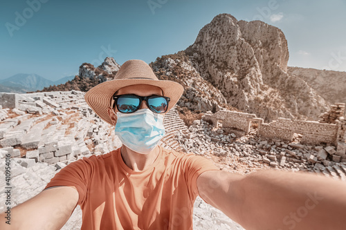 A male traveler wears a medical mask and sunglasses with a hat and takes a selfie against the ruins of the ancient amphitheater of Termessos. Quarantine and covid-19 concept