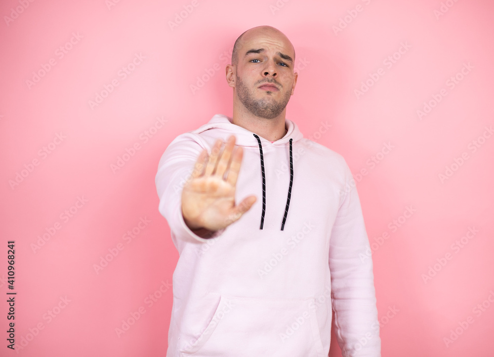 Young bald man wearing casual sweatshirt over pink isolated background serious and doing stop sing with palm of the hand. Young bald man wearing casual sweatshirt over pink isolated background serious and doing stop sing with palm of the hand.