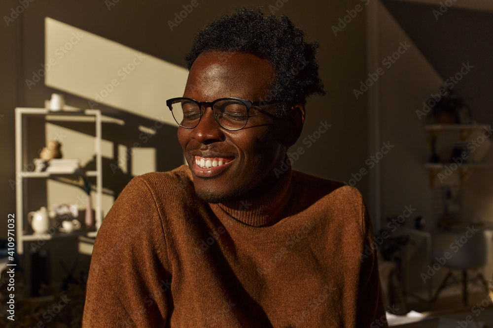 Fototapeta premium Portrait of stylish young Afro American man wearing brown sweater and glasses in black frame spending morning at home, enjoying sunny weather, sitting by window, smiling broadly, showing white teeth