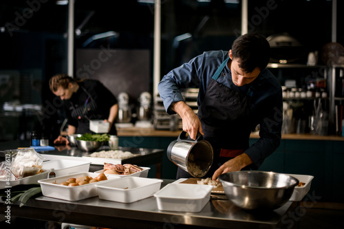 Fototapeta Naklejka Na Ścianę i Meble -  male chef in black apron puts ingredient from saucepan to cutting board