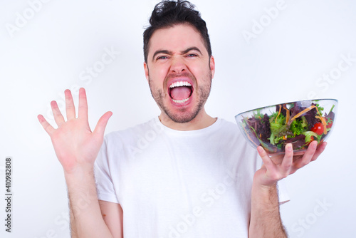 Optimistic Young handsome Caucasian man holding a salad bowl against white background raises palms from joy, happy to receive awesome present from someone, shouts loudly, Excited model screaming.