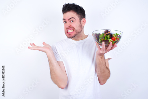 Clueless Young handsome Caucasian man holding a salad bowl against white background shrugs shoulders with hesitation, faces doubtful situation, spreads palms, Hard decision