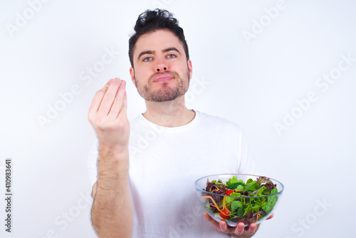 Young handsome Caucasian man holding a salad bowl against white background angry gesturing typical Italian gesture with hand, looking to camera