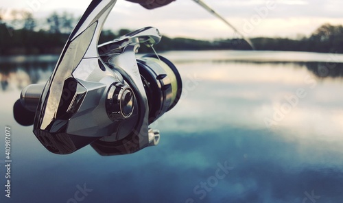 Fototapeta Naklejka Na Ścianę i Meble -  Fishing rod and spinning reel closeup with a lake in the background The concept of rural getaway. Article about fishing day.