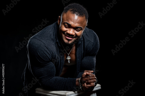 Handsome smiling muscular man in black shirt poses to the camera and leans on high bar chair with elbows. Black studio background.