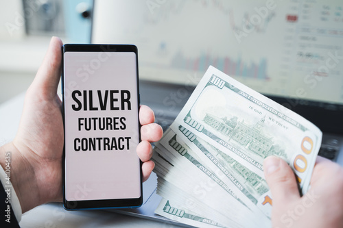 Mobile phone with text: silver futures contract and money, dollars in hand. Financial trading. Investment in precious metals. Playing on stock exchange, market manipulation.Losses, crisis.Short float photo