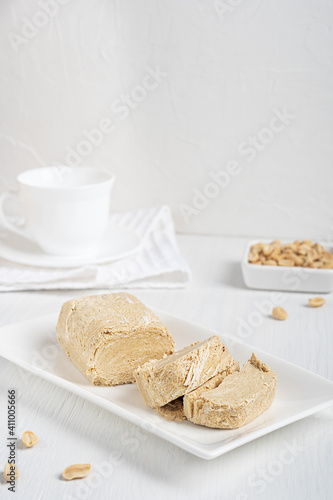 Side view of halva, also halvah or halwa is West Asia sweet dessert made of peanut oily seeds grinded to a paste and mixed with sugar syrup served on white plate on wooden table with towel and cup