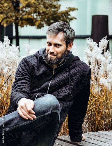 Handsome middle-aged Caucasian man portraited in the city, sitting on a bench with grey and black hair having a friendly look on his face, wearing a blue jacket and jeans. Fall colourful background.