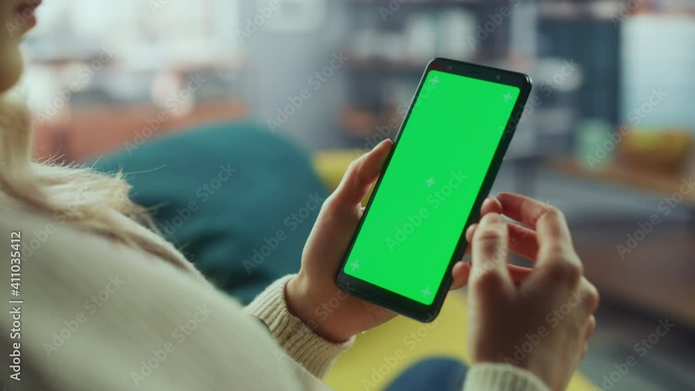 Close Up of an Authentic Female Using Smartphone with Green Screen Mock Up Display at Home Living Room while Lying on a Couch Sofa. She's Browsing the Internet and Checking Videos on Social Networks. Close Up of an Authentic Female Using Smartphone with Green Screen Mock Up Display at Home Living Room while Lying on a Couch Sofa. She's Browsing the Internet and Checking Videos on Social Networks.