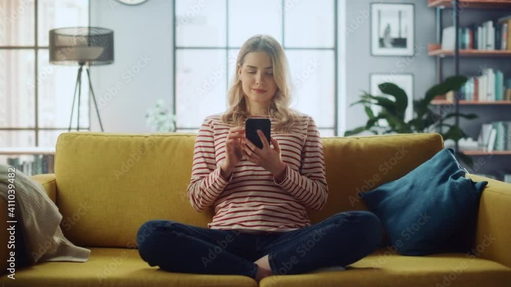 Beautiful Caucasian Female Using Smartphone in Stylish Living Room while Sitting on a Cozy Couch Sofa. Young Woman Resting at Home, Browsing Internet, Using Social Networks, Having Fun in Flat. Beautiful Caucasian Female Using Smartphone in Stylish Living Room while Sitting on a Cozy Couch Sofa. Young Woman Resting at Home, Browsing Internet, Using Social Networks, Having Fun in Flat.