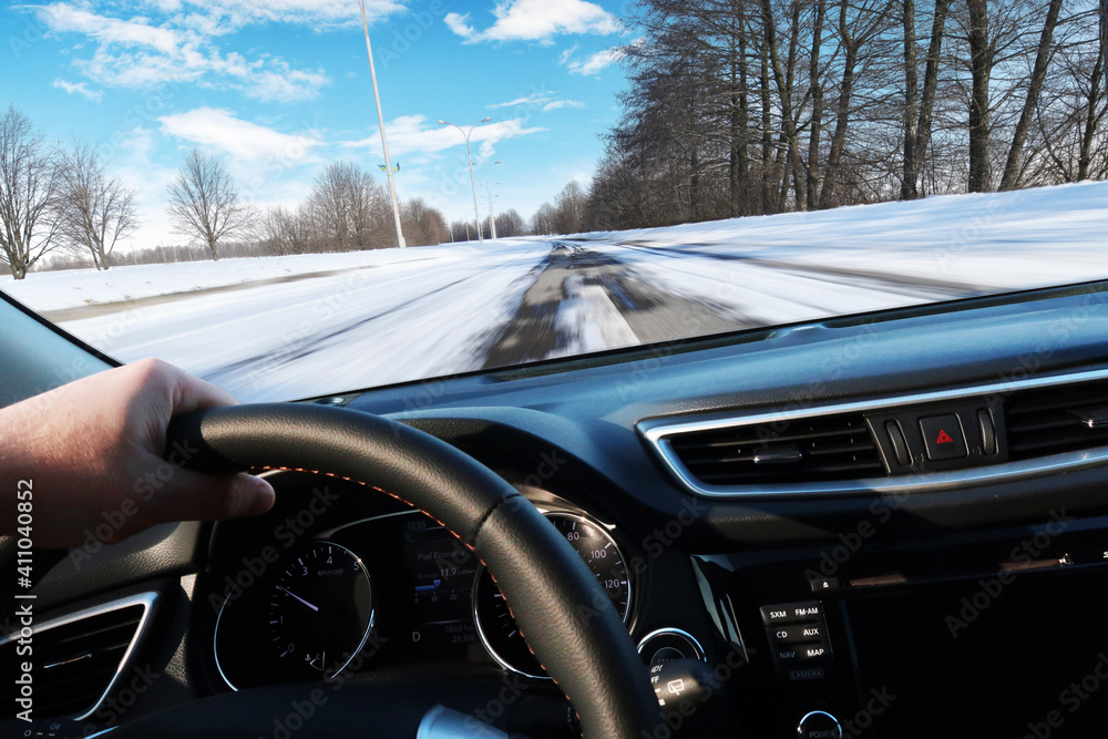 Car dashboard with driver's hand on the steering wheel with winter road in motion against a sky with clouds Car dashboard with driver's hand on the steering wheel with winter road in motion against a sky with clouds