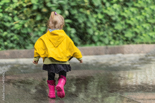 A happy little girl in a yellow raincoat and pink rubber boots walking in the rain on the street alone. Nature, outdoors. Childhood concept. Universal Children's Day.