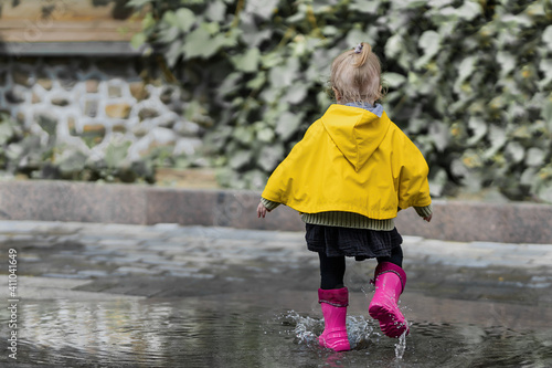 Fototapeta Naklejka Na Ścianę i Meble -  A nice little girl in a yellow raincoat and pink rubber boots jumps on puddles with splashes and rejoices. Park, nature, outdoors. Summer time. Universal Children's Day. babyhood