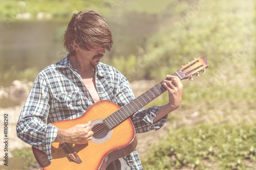  Adult male musician in a plaid shirt is playing the guitar on nature. Bright, sunny day, summer, outdoors. Close-up portrait. Retro concept.