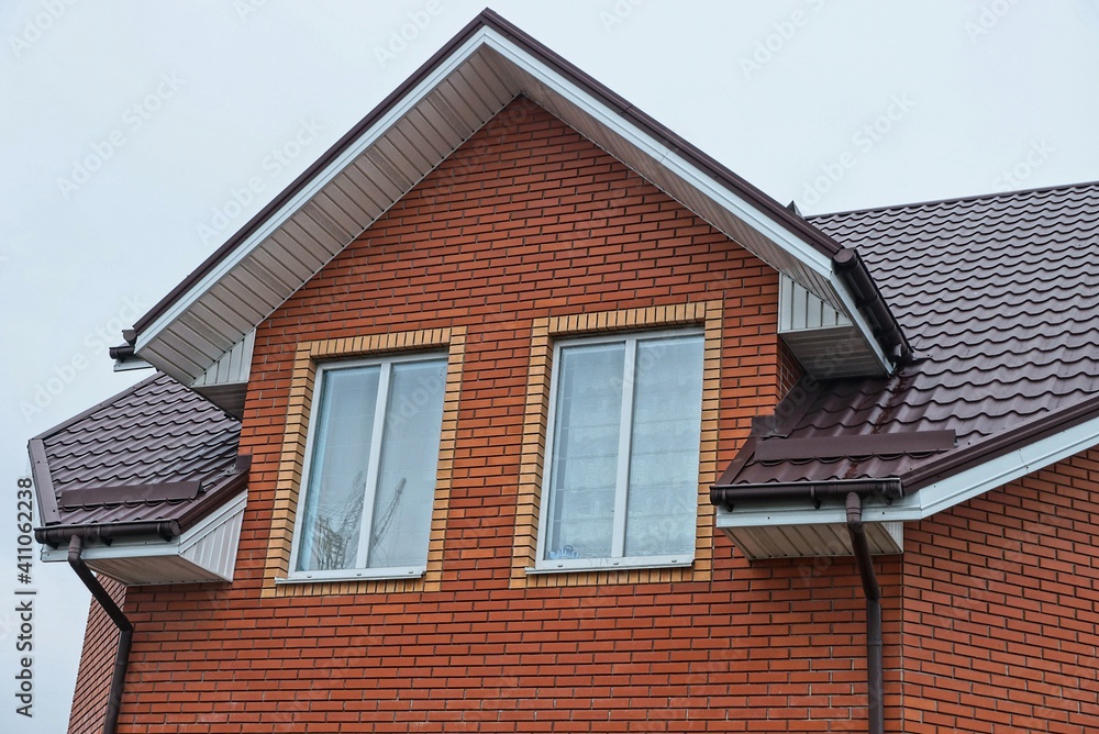  attic of a red brick private house with a two windows under a brown tiled roof against a gray sky