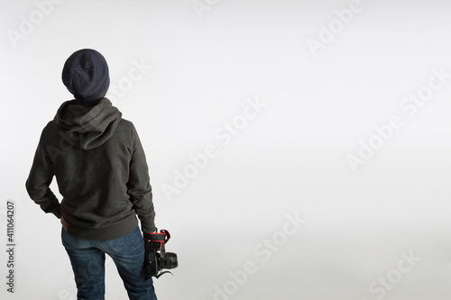 A teenager with a SLR camera on his back stands in a hoodie and a hat on a blue background.
