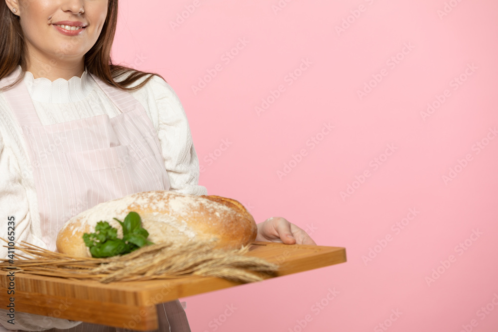 A young attractive saleswoman from a bakery shop holds a loaf of wheat flour bread and a pinch of fresh green herb leaves and rye ears on a wooden tray. The whole on a uniform light pink background. A young attractive saleswoman from a bakery shop holds a loaf of wheat flour bread and a pinch of fresh green herb leaves and rye ears on a wooden tray. The whole on a uniform light pink background.