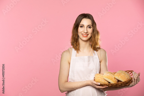 An attractive saleswoman holds three freshly baked rolls in a wicker basket. Seamless light pink background.