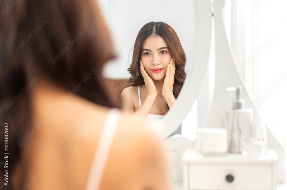 Smiling of young beautiful pretty asian woman clean fresh healthy white skin looking at mirror.asian girl touching on her face with hand and applying cream at home.spa and beauty concept Smiling of young beautiful pretty asian woman clean fresh healthy white skin looking at mirror.asian girl touching on her face with hand and applying cream at home.spa and beauty concept