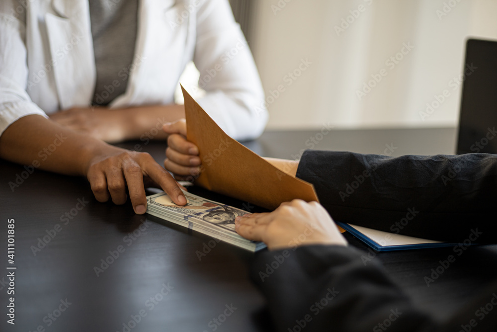Two women holding a dollar under a brown envelope. A businesswoman who is handing over a lot of dollar bills to a business partner as a bribe, bribery is an illegal act. Corruption concept. Two women holding a dollar under a brown envelope. A businesswoman who is handing over a lot of dollar bills to a business partner as a bribe, bribery is an illegal act. Corruption concept.