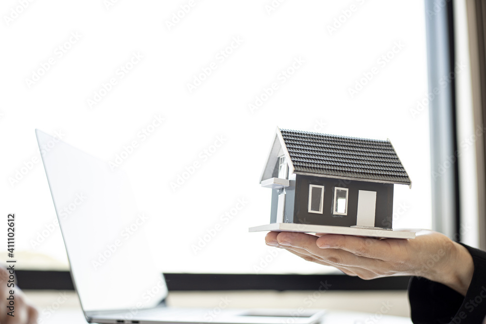 A female home salesman holds a gray miniature house model to describe the details of the house to the customer who will purchase a house on the project. Real estate trading ideas. A female home salesman holds a gray miniature house model to describe the details of the house to the customer who will purchase a house on the project. Real estate trading ideas.