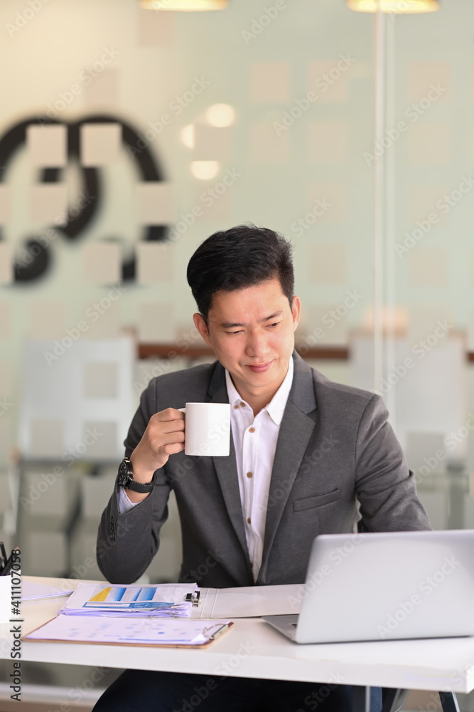 Portrait os smart businessman holding coffee cup and scrolling through online information on laptop computer at modern office. Portrait os smart businessman holding coffee cup and scrolling through online information on laptop computer at modern office.