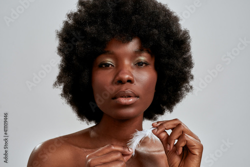 Portrait of beautiful african american young woman with afro hair looking at camera, holding white feather, posing isolated over gray background photo