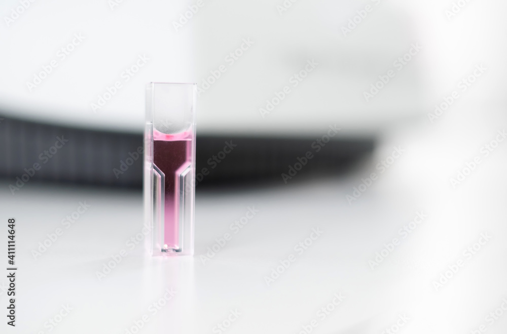 Plastic cuvette contains a pink solution sample on the table with copy space in the laboratory. Background is blurry Ultraviolet (UV) spectroscopy machine. Research and education concept. Plastic cuvette contains a pink solution sample on the table with copy space in the laboratory. Background is blurry Ultraviolet (UV) spectroscopy machine. Research and education concept.