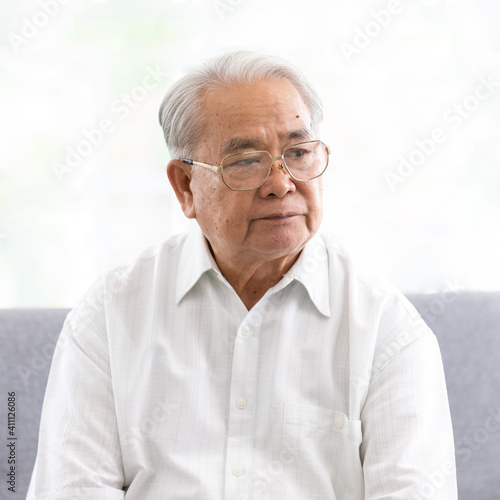 Portrait of old senior asian man wearing eyesglasses and white shirt sitting on sofa.