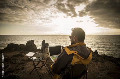 Digital nomad people at work outdoor with computer laptop and 5g internet connection in roaming phone hotspot - man travel and work alone sitting and enjoying the sunset on the ocean coast