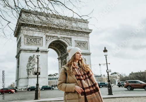 Young beautiful fashionable woman tourist in  white hat and scarf on background of  famous Arc de Triomphe or Triumphal arch. Winter or autumn in Europe. Paris, France.