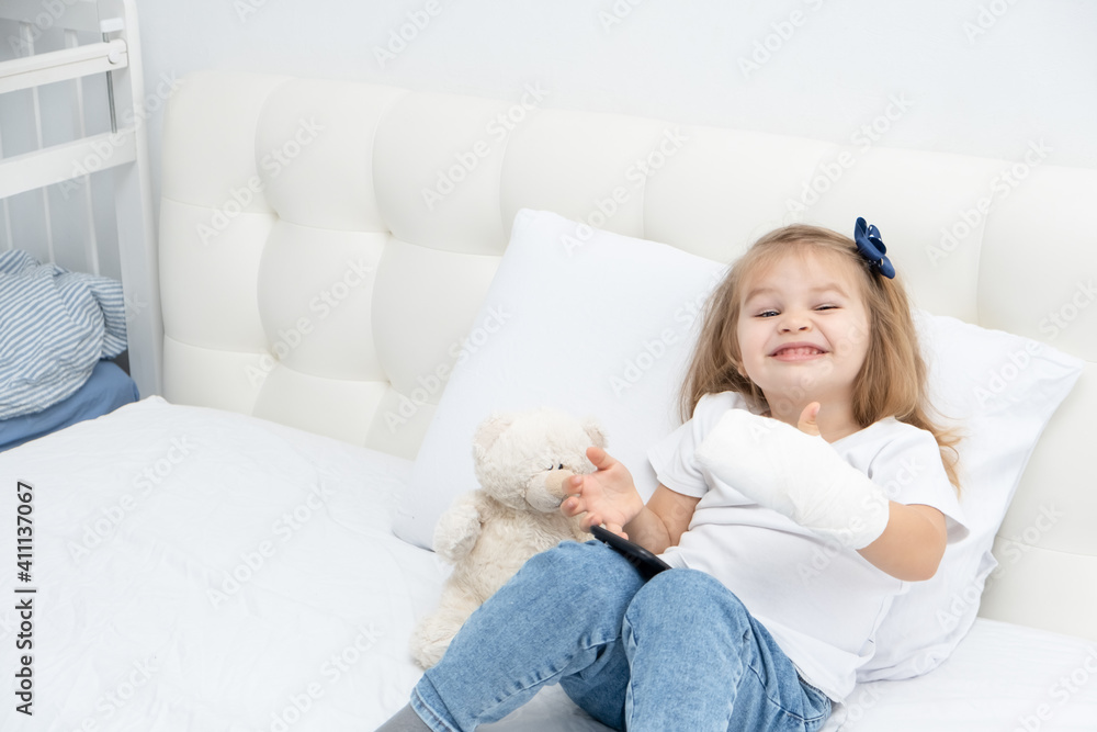 Little girl with hand in plaster sitting in bed showing the class, using smartphone. Little girl with hand in plaster sitting in bed showing the class, using smartphone.