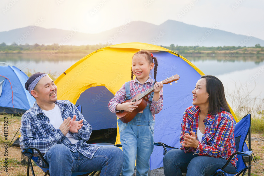 Happy little asian girl playing ukulele and her parents clapping hands at camping site.